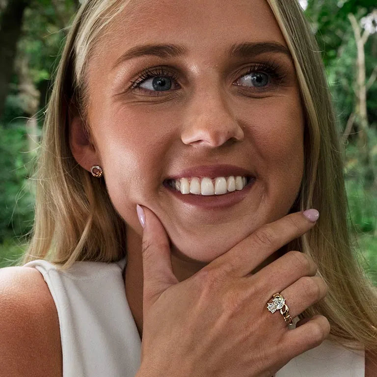 A close-up lifestyle shot of a smiling blonde woman outdoors, wearing a gold ring with a clear gemstone on her ring finger and a matching stud earring. The background is a soft-focus green forest. 14K Gold | Claddagh Ring with Lab Diamonds