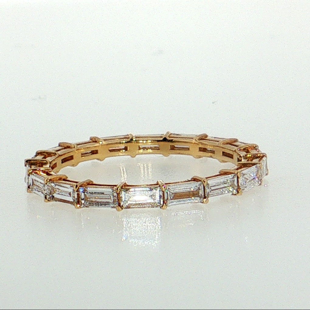A gold eternity ring featuring baguette-cut clear stones, photographed on a reflective white surface against a bright white background.