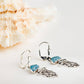 A pair of silver leverback earrings with blue crystal accents and dangling silver wing charms, displayed on a light surface next to a seashell.