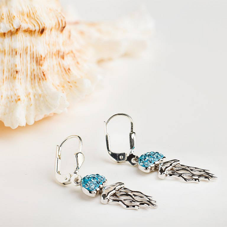A pair of silver leverback earrings with blue crystal accents and dangling silver wing charms, displayed on a light surface next to a seashell.