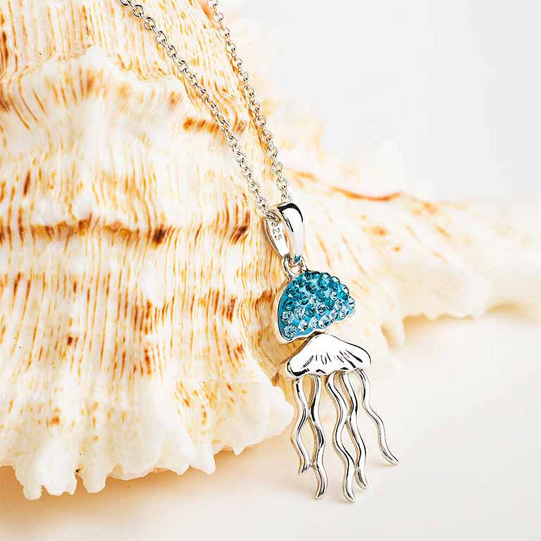 A silver jellyfish pendant necklace with a bright blue rhinestone-studded top, displayed on a large beige seashell against a clean white background. Ocean | Deep Blue Sterling Silver Aqua Crystal Jellyfish Necklace