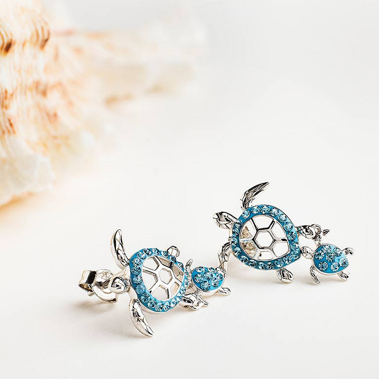 A pair of silver turtle stud earrings decorated with bright blue rhinestones, displayed on a clean white surface with a soft-focus seashell in the background.