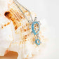 Silver-toned turtle pendant necklace with light blue rhinestone detailing, displayed on a large white seashell against a bright white background.