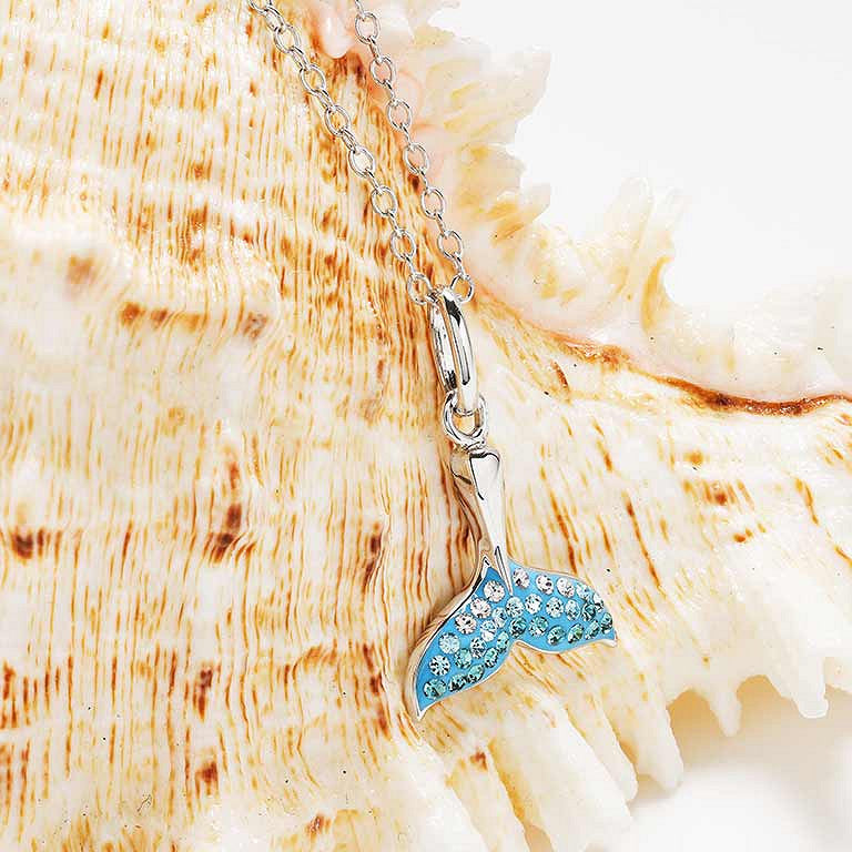 A silver-toned mermaid tail pendant necklace with light blue enamel and clear rhinestones, displayed on a large beige seashell. Ocean | Deep Blue Sterling Silver Aqua Crystal Whale Tail mini necklace