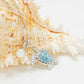 A silver-toned turtle pendant necklace with a light blue rhinestone-embellished shell and a dangling white pearl, displayed on a large seashell against a white background.