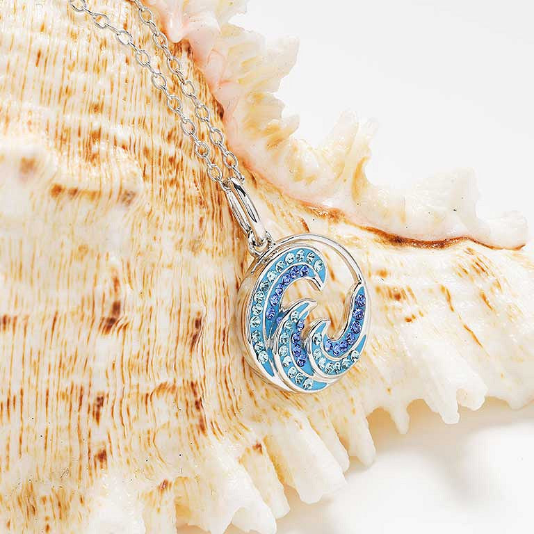 A silver wave pendant necklace with blue rhinestone detailing, displayed on a large beige seashell against a white background.