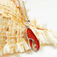 A lobster claw pendant necklace with textured red enamel and silver-tone metal, resting on a large seashell against a white background.