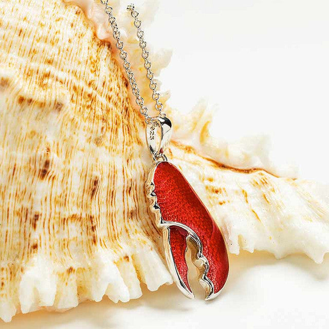A lobster claw pendant necklace with textured red enamel and silver-tone metal, resting on a large seashell against a white background.