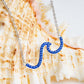 A silver wave-shaped pendant necklace paved with small blue stones, draped over a large seashell. Ocean | Deep Blue Sterling Silver Wave Necklace with Sapphire Blue crystals