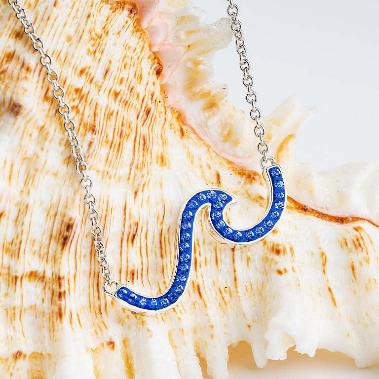 A silver wave-shaped pendant necklace paved with small blue stones, draped over a large seashell. Ocean | Deep Blue Sterling Silver Wave Necklace with Sapphire Blue crystals