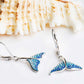 A pair of dangling earrings shaped like mermaid tails, decorated with sparkling blue rhinestones and silver-tone metal hooks, displayed on a white surface with a seashell in the background.