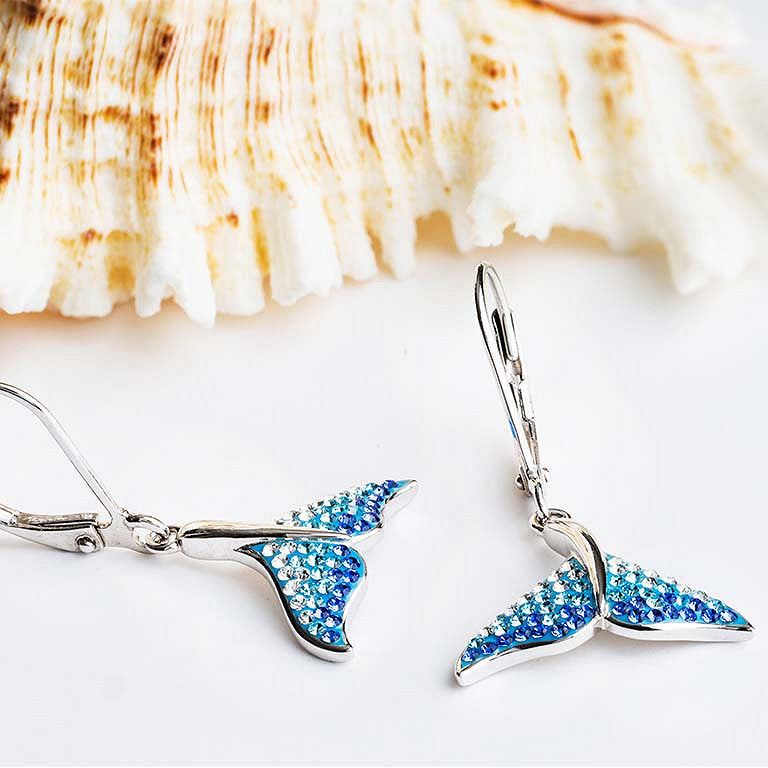 A pair of dangling earrings shaped like mermaid tails, decorated with sparkling blue rhinestones and silver-tone metal hooks, displayed on a white surface with a seashell in the background.