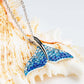 A mermaid tail pendant necklace with blue rhinestone-embellished wings and a silver chain, displayed on a large seashell.