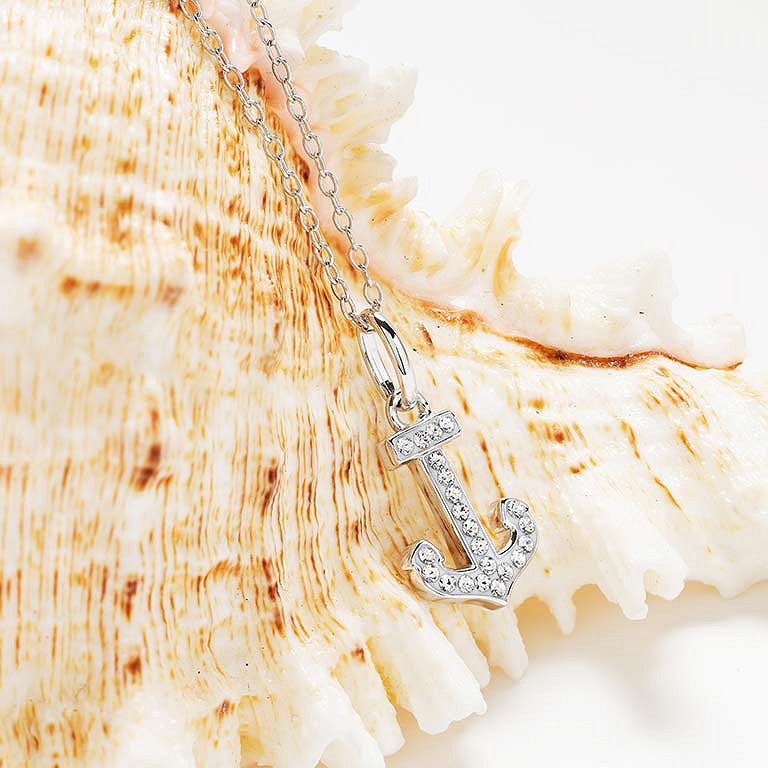 A silver anchor pendant necklace adorned with small clear rhinestones, resting on a large beige seashell against a bright white background. 