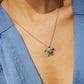 A silver-toned necklace with a turtle pendant featuring green and white stone detailing, worn by a person wearing a blue top.