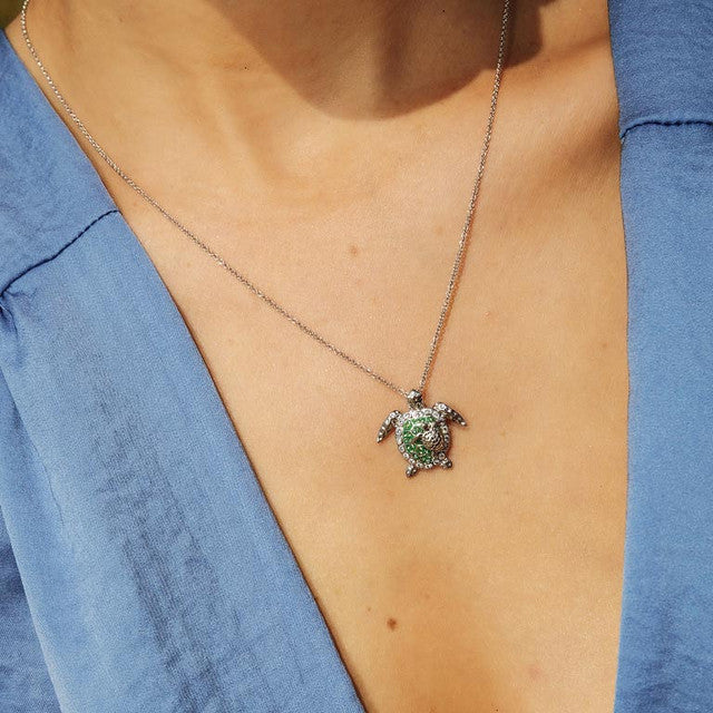 A silver-toned necklace with a turtle pendant featuring green and white stone detailing, worn by a person wearing a blue top.