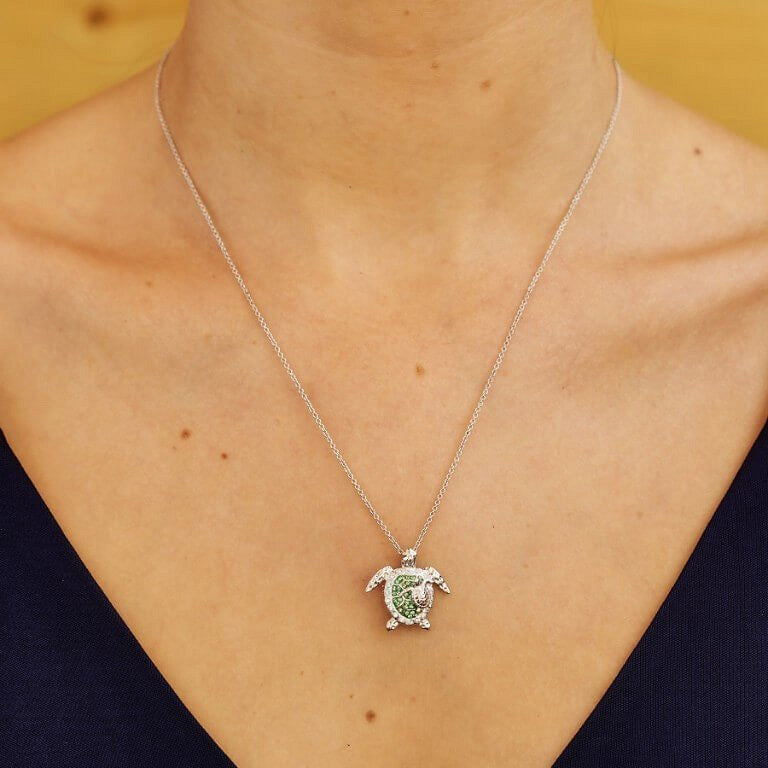 A silver-toned sea turtle pendant necklace with a green center, worn on a model's neck over a dark navy V-neck top.