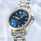 A Seiko women's watch featuring a silver stainless steel link bracelet, a blue dial with gold-tone hands and hour markers, and a gold bezel accent, set against a light blue water-textured background.